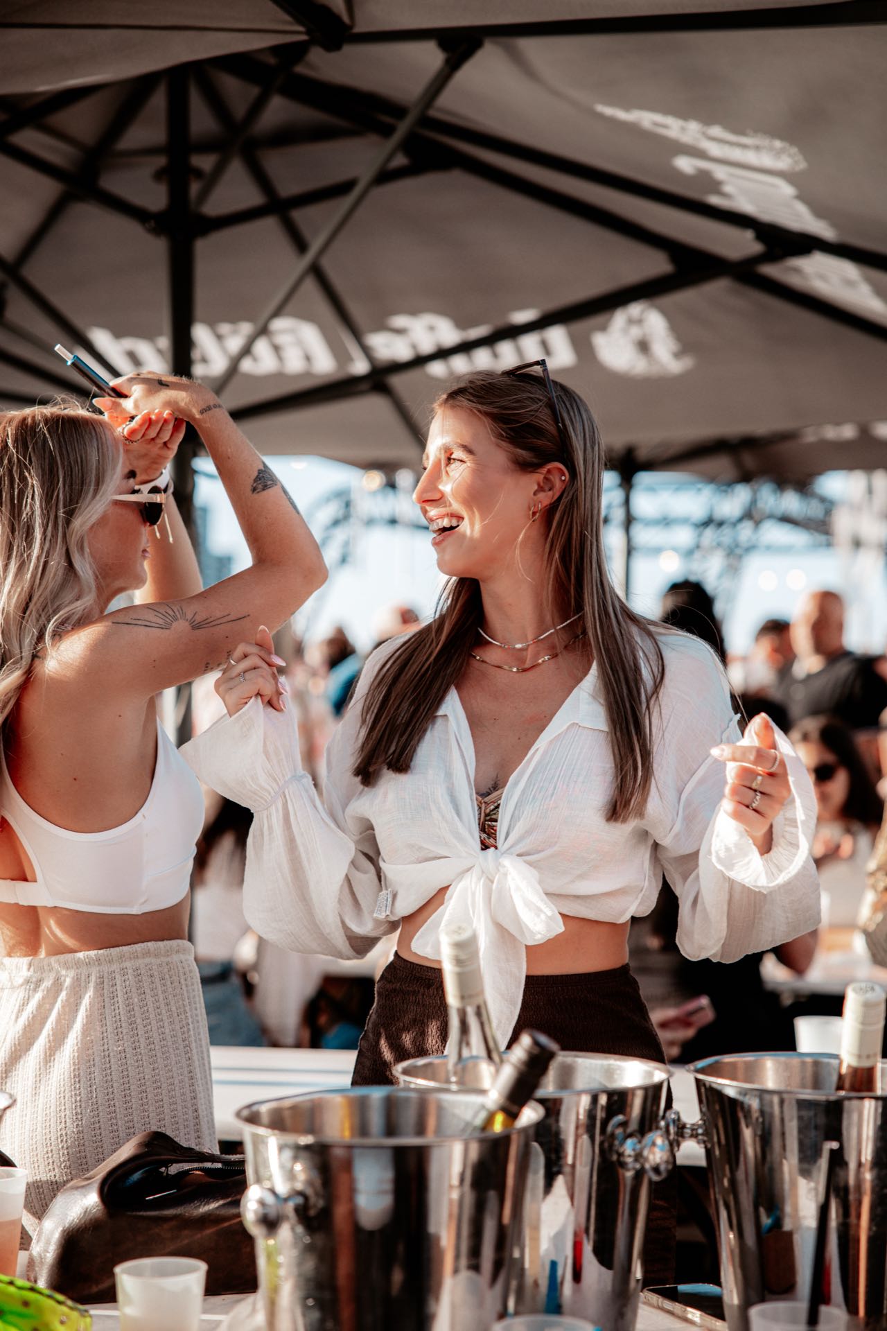 Zwei Freundinnen halten sich an den Händen und lachen sich an, unter Sonnenschirmen beim Golden Flame Open-Air an den Rheinterrassen Köln