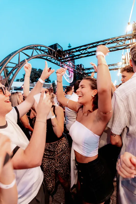 Freunde tanzen zur blauen Stunde mit erhobenen Armen beim Open Air in Köln, Lichterketten im Hintergrund