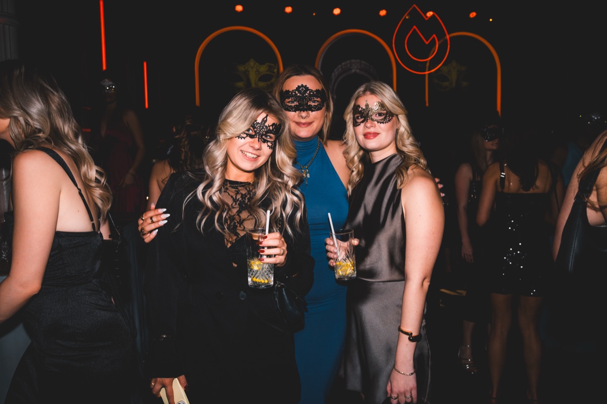 Drei Freundinnen mit Masken vor dem FAYA-Logo beim Golden Mask Maskenball in der Flora Köln
