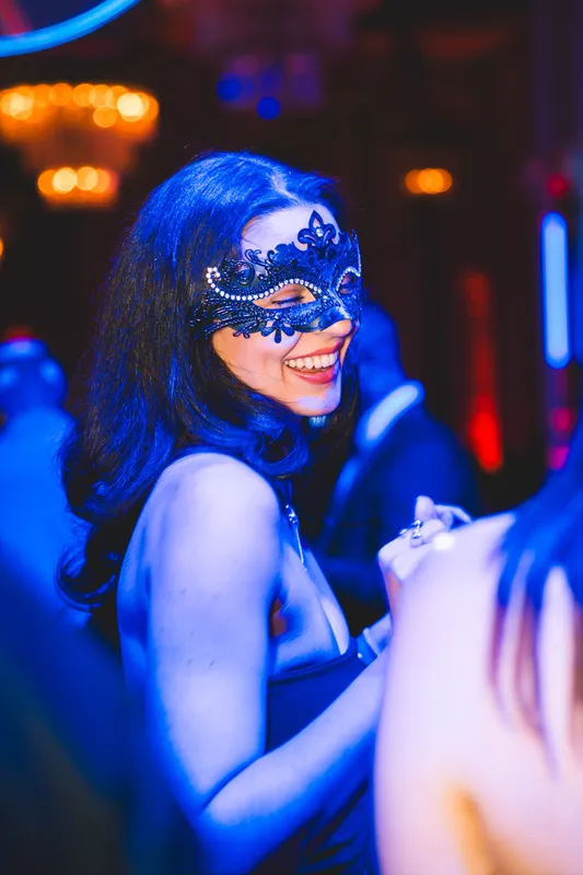 Frau mit langen dunklen Wellen und schwarzer Spitzenmaske lacht im blauen Licht beim Golden Mask