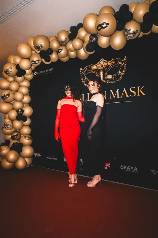 Fotowand mit großer goldener Maske und Ballonbogen in Gold und Schwarz, davor zwei Gäste in Rot und Schwarz beim Golden Mask