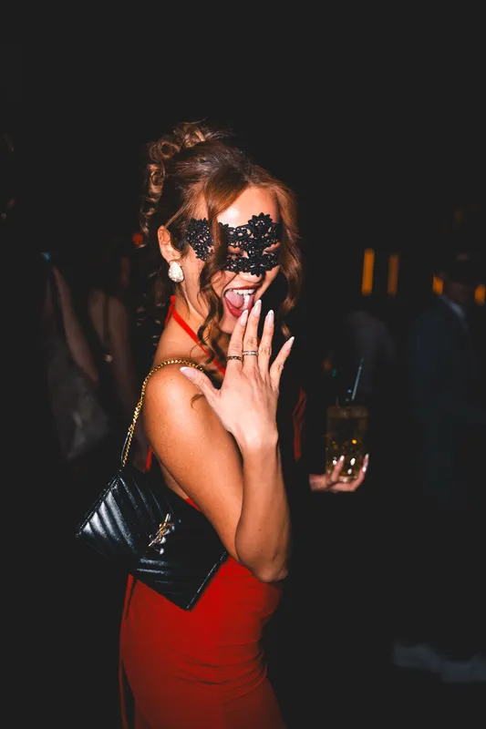 Lachende Frau mit lockerer Hochsteckfrisur, schwarzer Spitzenmaske und rotem Lippenstift im roten Kleid beim Maskenball