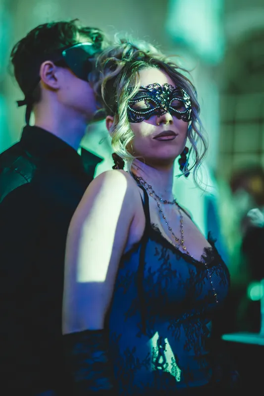 Frau mit schwarzer Spitzenmaske und lockiger Hochsteckfrisur, hinter ihr ein Gast mit schwarzer Maske, beim Golden Mask