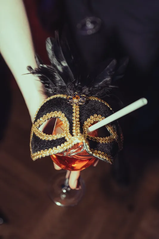 Hand hält ein Glas, darauf eine schwarz-goldene Maske mit Federn, beim Golden Mask Maskenball
