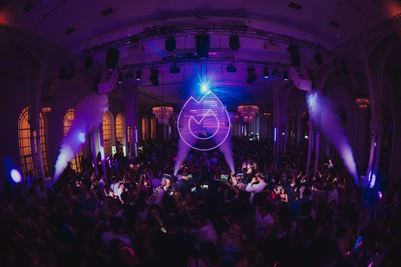 Festsaal in violettem Licht mit leuchtendem Flammen-Logo und voller Tanzfläche bei Golden Mask