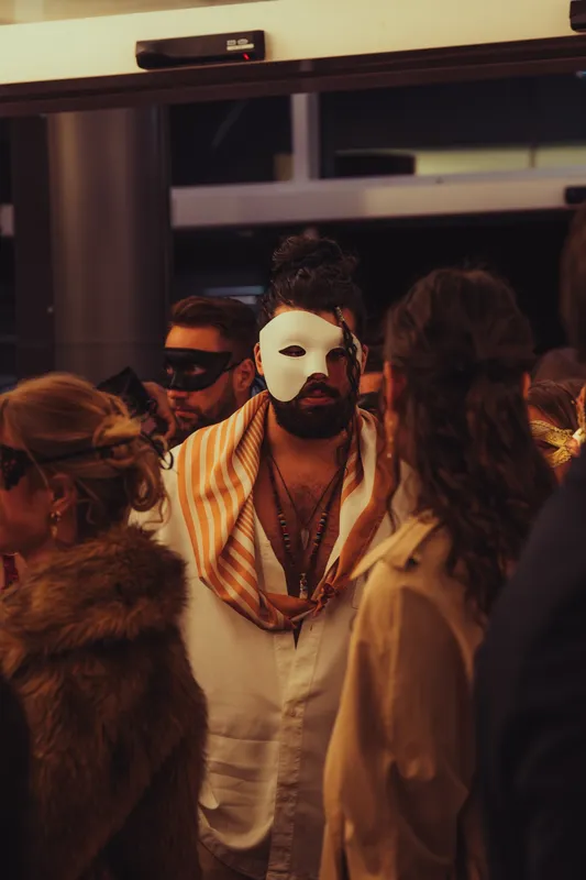 Gast mit weißer Maske, die das halbe Gesicht bedeckt, und gestreiftem Schal beim Maskenball Golden Mask in Köln