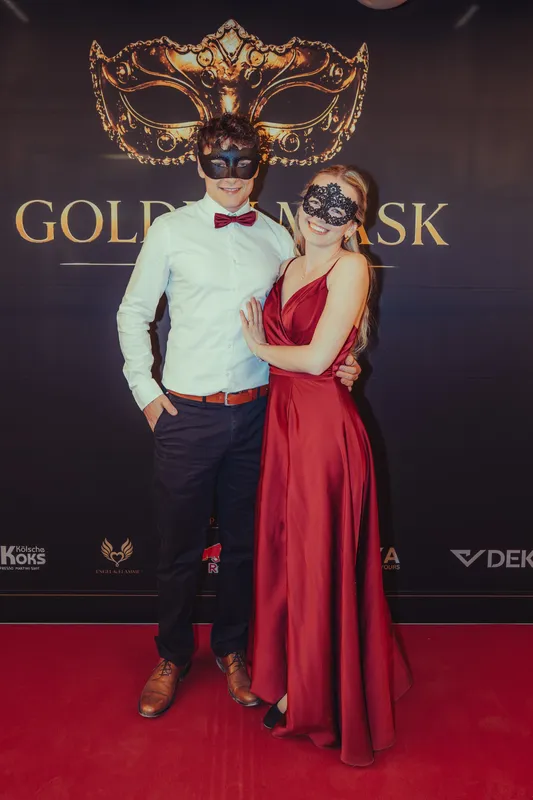 Paar beim Maskenball: er in weißem Hemd mit roter Fliege, sie im roten Satinkleid, beide mit schwarzen Masken