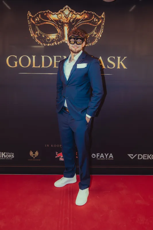 Mann im blauen Anzug mit weißem Hemd, weißen Sneakern und schwarzer Maske vor der Golden-Mask-Fotowand