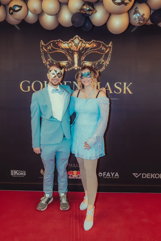Paar im hellblauen Partnerlook mit Masken vor der Golden-Mask-Fotowand mit goldener Maske in der Flora Köln