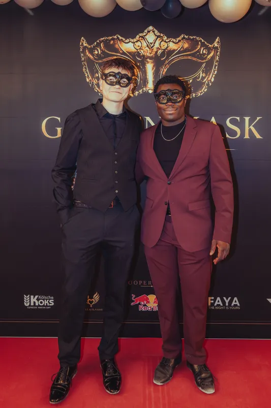 Zwei Männer mit schwarzen Masken, einer im dunklen Anzug mit Weste, einer im bordeauxfarbenen Anzug, auf dem roten Teppich beim Golden Mask
