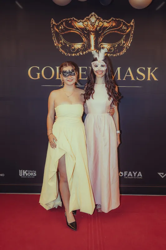 Zwei Frauen in langen Kleidern in Pastellgelb und Creme mit Masken vor der Golden-Mask-Fotowand