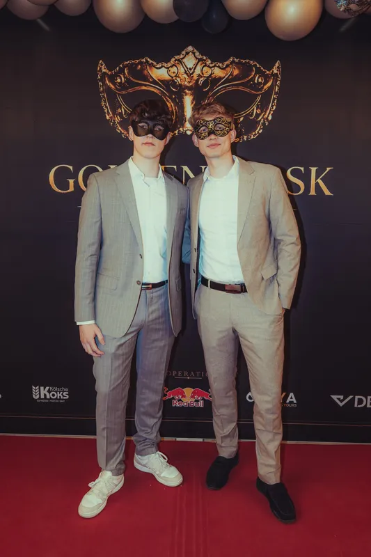 Zwei Männer in hellgrauen und beigen Anzügen mit weißen Hemden und schwarzen Masken vor der Fotowand beim Golden Mask in Köln