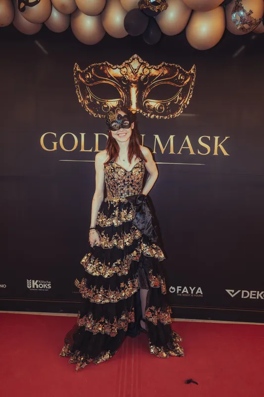 Frau in bodenlanger schwarzer Spitzenrobe mit Rüschen und Maske vor der goldenen Fotowand beim Golden Mask in Köln