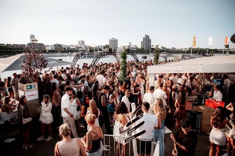 A busy terrace with guests in summer outfits overlooking the Rhine and the Cologne skyline