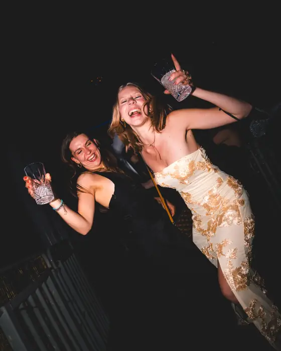 Zwei Freundinnen lachen ausgelassen, links im schwarzen Kleid, rechts im cremefarbenen Kleid mit goldener Stickerei, beim Golden Mask in der Flora Köln