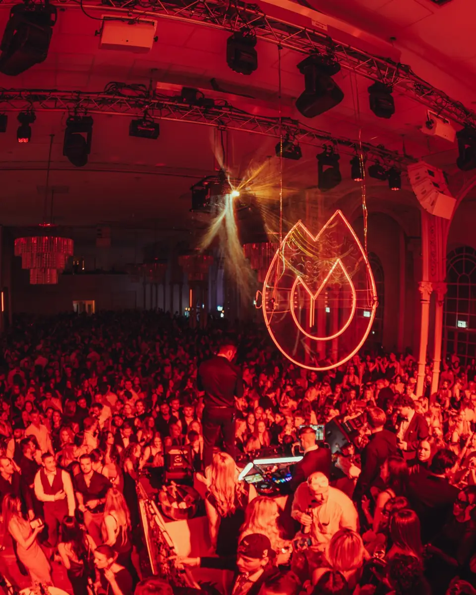 Volle feestzaal in rood licht met het verlichte vlammenlogo tijdens Golden Mask in de Flora in Keulen