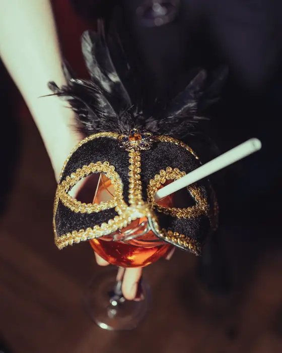 Hand hält eine goldene Maske mit Federn und ein Glas beim Golden Mask Maskenball