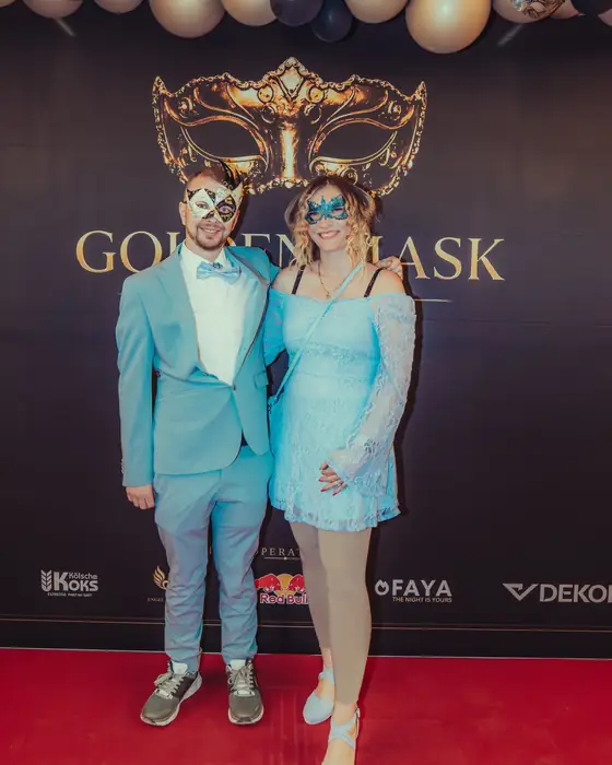 Paar im Partnerlook vor der Golden-Mask-Fotowand: er im hellblauen Anzug mit Fliege, sie im hellblauen Spitzenkleid, beide mit hellen Masken