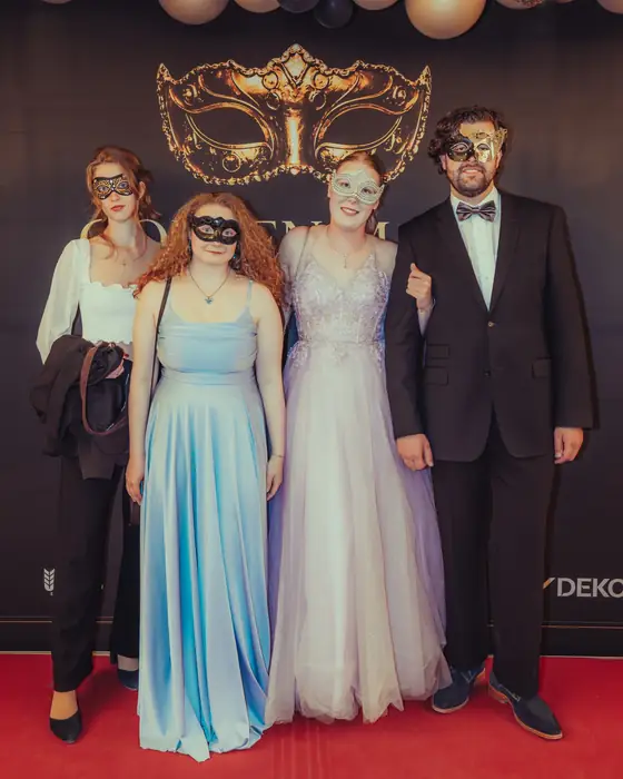 Vier Gäste mit Masken vor der Golden-Mask-Fotowand: zwei Frauen in langen Roben in Hellblau und Flieder, eine Frau in Schwarz und ein Mann im Anzug mit Fliege