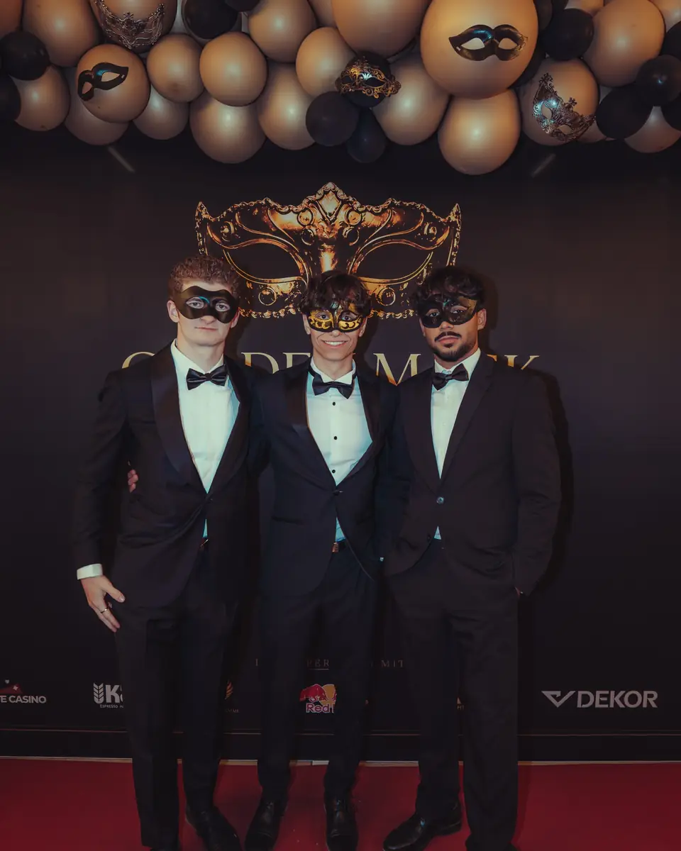 Drei Männer im schwarzen Smoking mit Fliege und schwarzen Masken vor der Golden-Mask-Fotowand mit goldenen Ballons in der Flora Köln