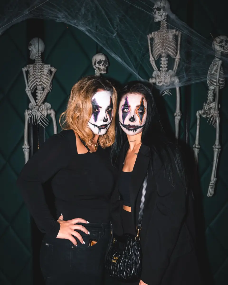 Zwei Freundinnen mit weiß geschminkten Gesichtern und schwarzen Augen vor einer Tür mit Skeletten bei Spooky Flame