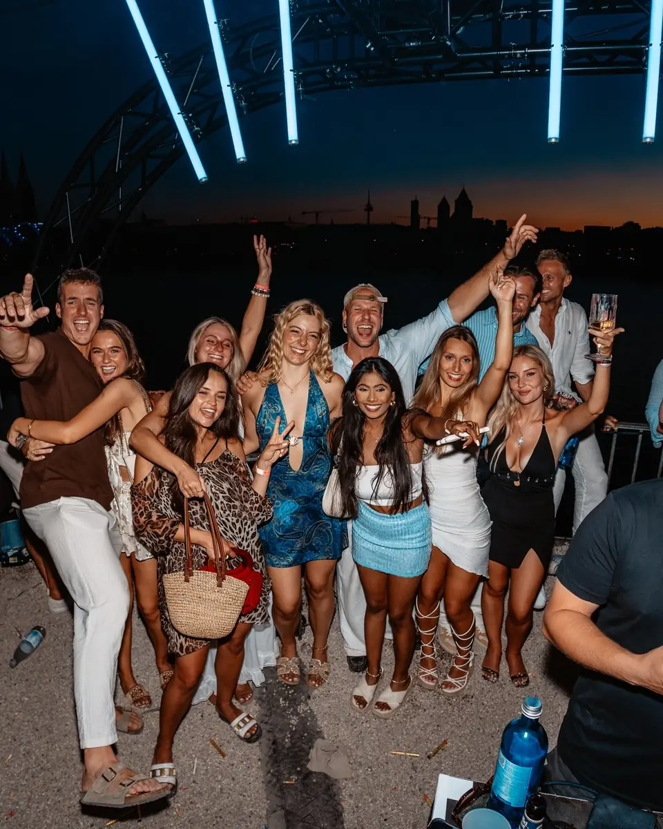 A group of friends celebrating with their arms in the air at blue hour after sunset at the Rheinterrassen in Cologne
