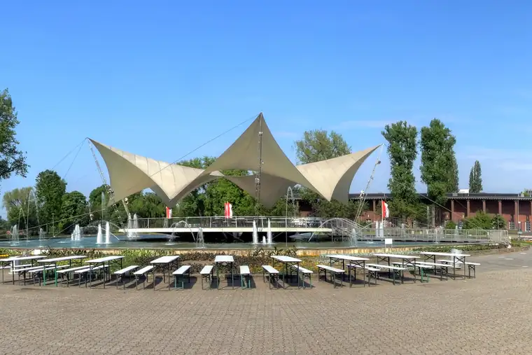 Das weiße Sternwellenzelt von Frei Otto am Tanzbrunnen Köln über dem Brunnen, davor Tische und Bäume im Rheinpark
