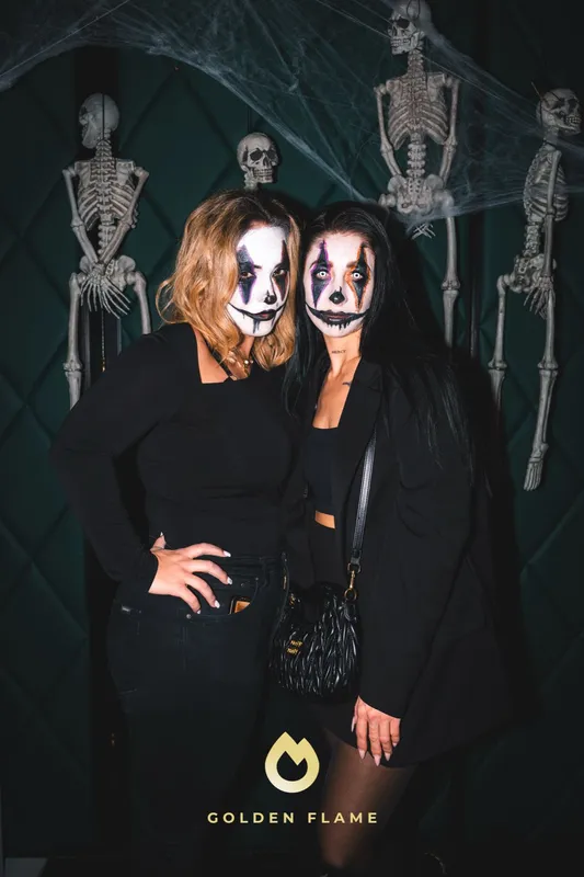 Zwei Freundinnen mit weiß geschminkten Gesichtern und schwarzen Augen vor einer Tür mit Skeletten bei Spooky Flame