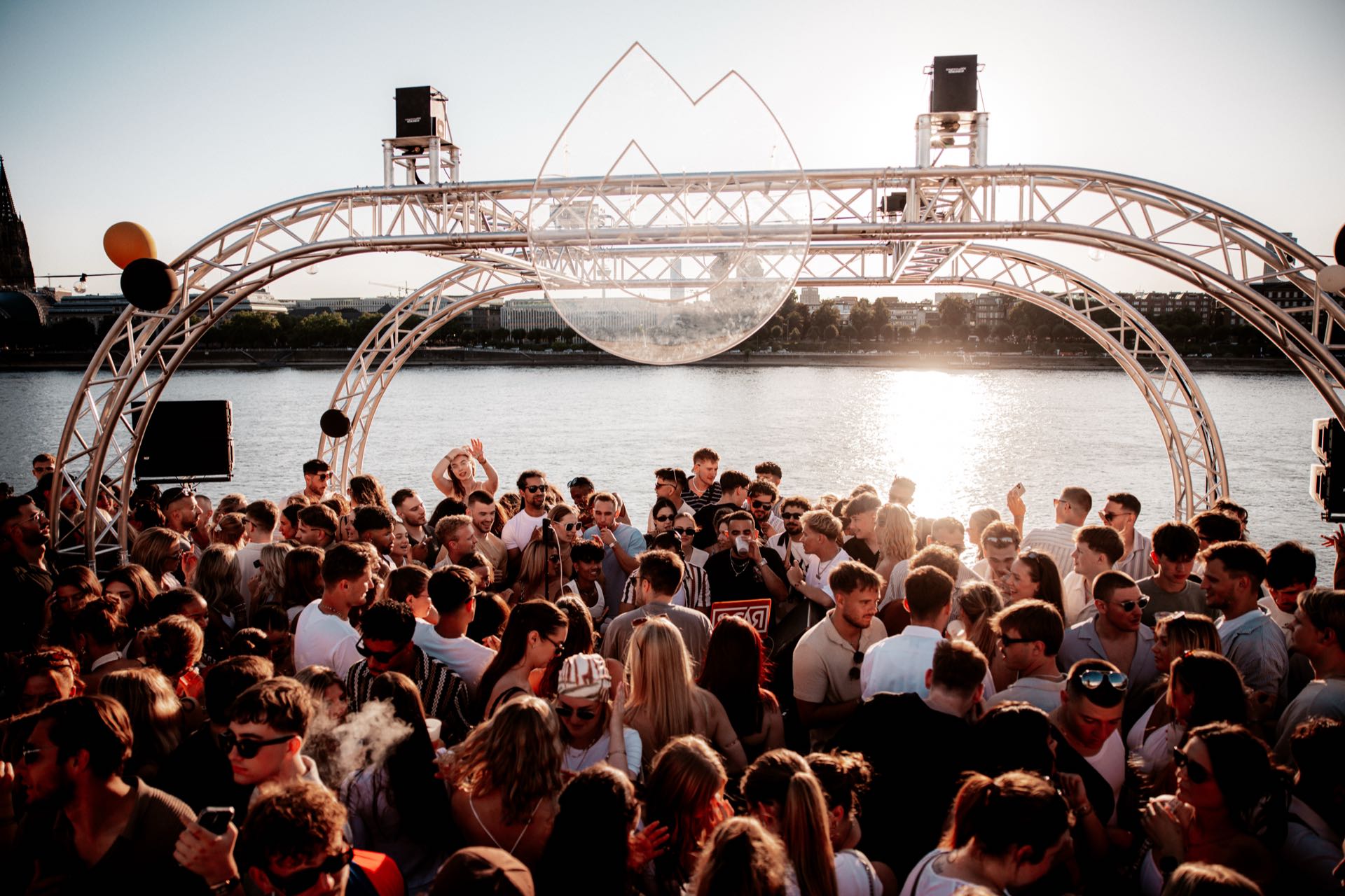 Weite Sicht auf das Golden Flame Open-Air am Rheinufer in Köln bei Sonnenuntergang, die Tanzfläche voller Menschen