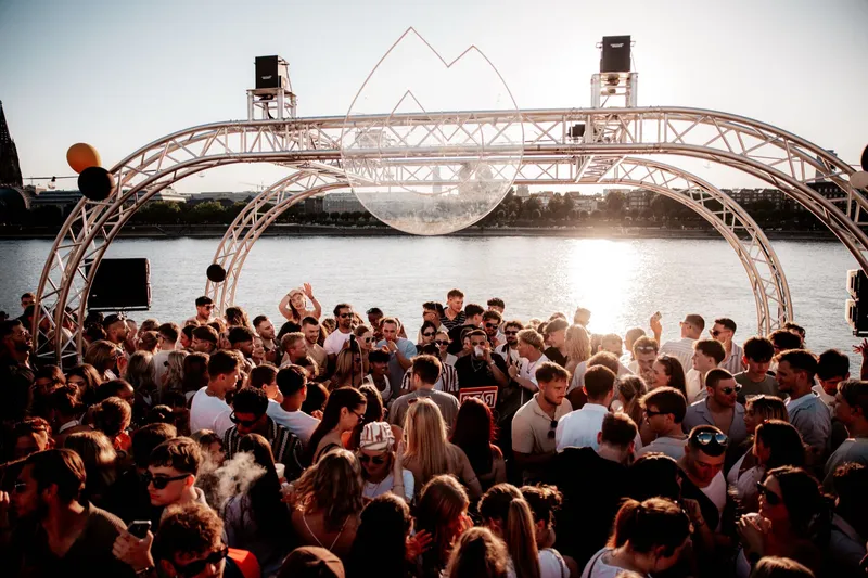 Weite Sicht auf das Golden Flame Open-Air am Rheinufer in Köln bei Sonnenuntergang, die Tanzfläche voller Menschen